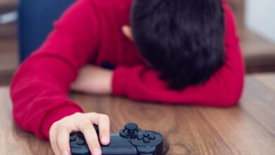 Niño con camisa roja de manga larga recostado en una mesa de madera con la cabeza en su brazo isquierdo escondiendo la cara. Su mano derecha aguanta un control de videojuegos.