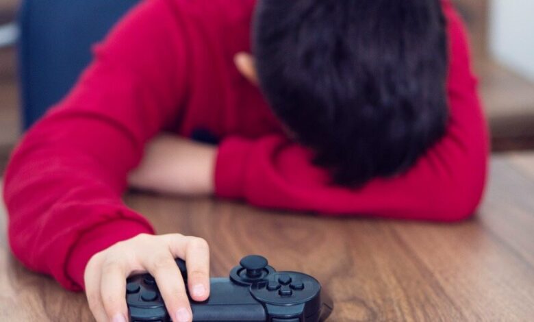 Niño con camisa roja de manga larga recostado en una mesa de madera con la cabeza en su brazo isquierdo escondiendo la cara. Su mano derecha aguanta un control de videojuegos.